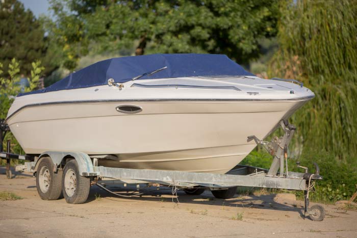 A white motorboat with a blue canvas cover sits securely on a dual-axle boat trailer, parked on a concrete surface with green trees in the background.
