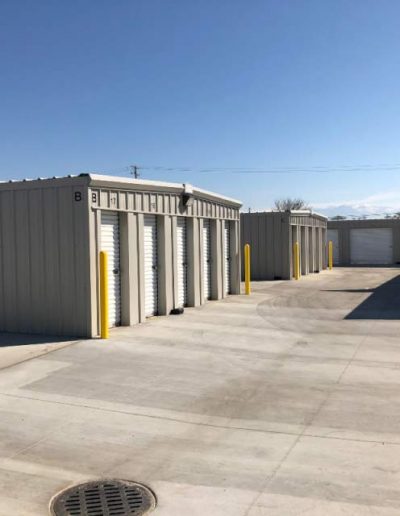 Wide outdoor view of a secure commercial self-storage facility complex with wide concrete driveways, multiple numbered storage buildings, and yellow protective bollards under a clear sunny sky.