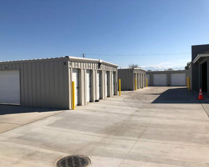 Wide-angle view of a professional commercial storage facility in Visalia, showing multiple rows of clean, tan metal storage buildings with secure white roll-up doors. The image features a large, paved concrete driveway that provides ample space for trucks and commercial vehicles to maneuver, highlighted by bright yellow safety bollards and a clear blue sky in the background.