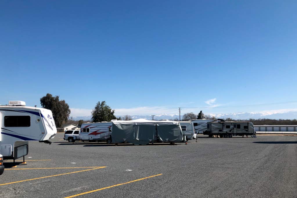 An expansive outdoor vehicle storage area featuring several parked RVs and travel trailers under a clear blue sky with a view of snow-capped mountains in the distance.