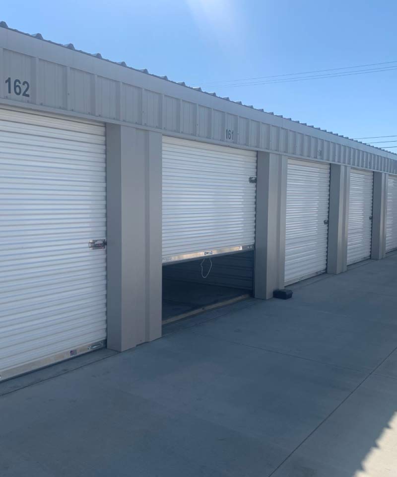 A row of well-maintained self-storage units with white roll-up doors and numbered headers, featuring a wide concrete driveway for easy vehicle access.