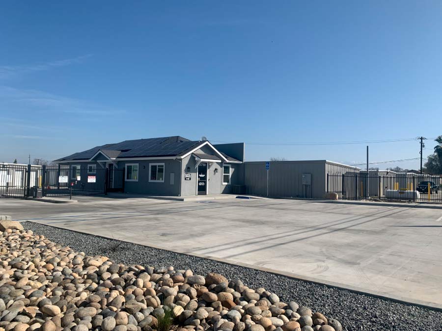 Exterior view of the Freedom Storage rental office, a modern gray building with solar panels on the roof, a clean concrete parking area, and a secure black perimeter fence.
